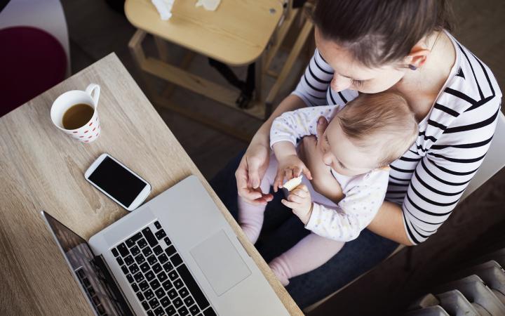 An image showing a woman holding a baby while sitting at a table that holds a laptop, mobile phone and cup of coffee.